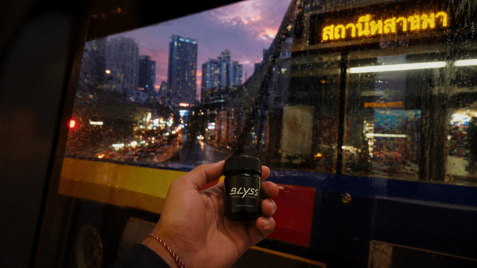 Hand holding BLYSS on a Bangkok bus at dusk, Thai signage and skyline visible through the window